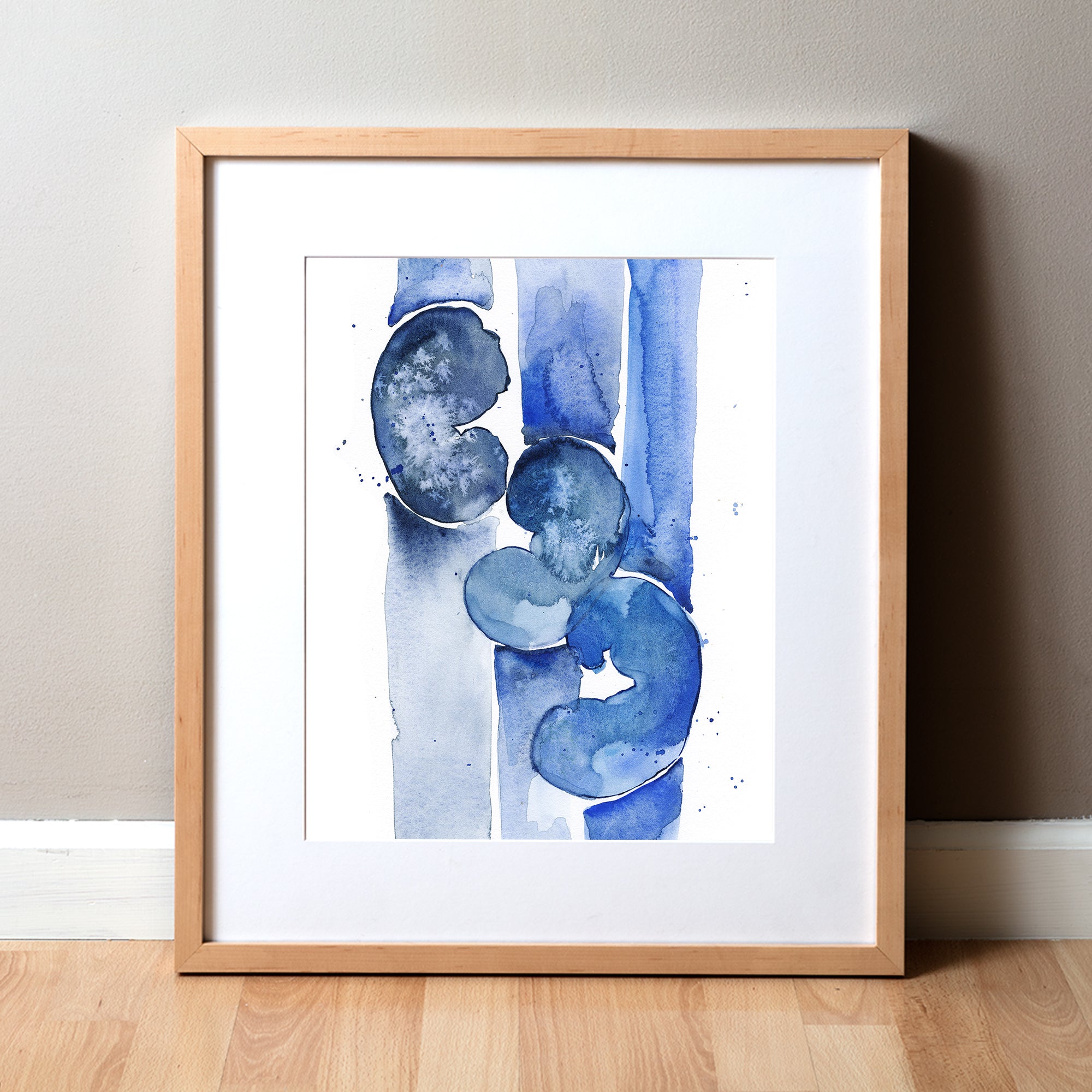 A watercolor painting of three kidneys in blues and navy. It is abstract, but still clearly kidneys to a nephrologist. The anatomy art print is in a natural light wood frame propped up on a beige wall and light wood floor.