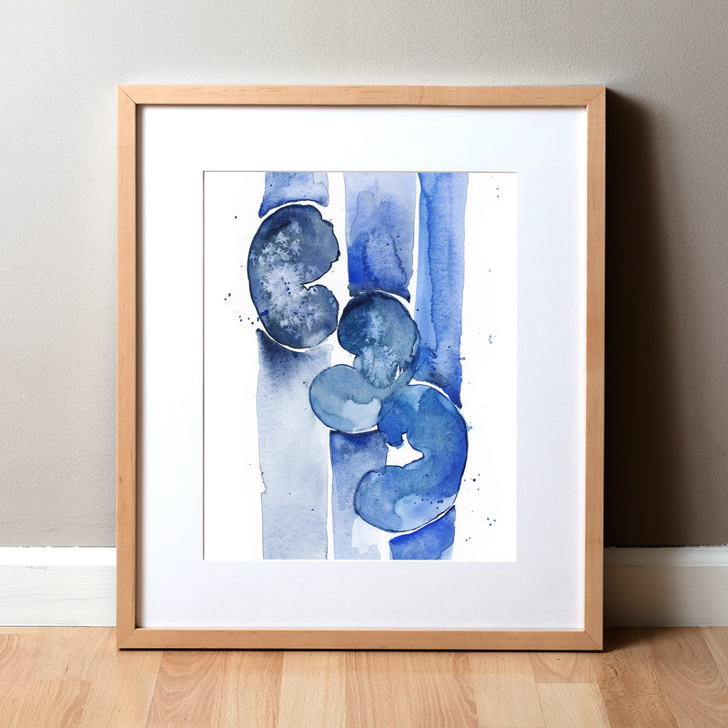 A watercolor painting of three kidneys in blues and navy. It is abstract, but still clearly kidneys to a nephrologist. The anatomy art print is in a natural light wood frame propped up on a beige wall and light wood floor.