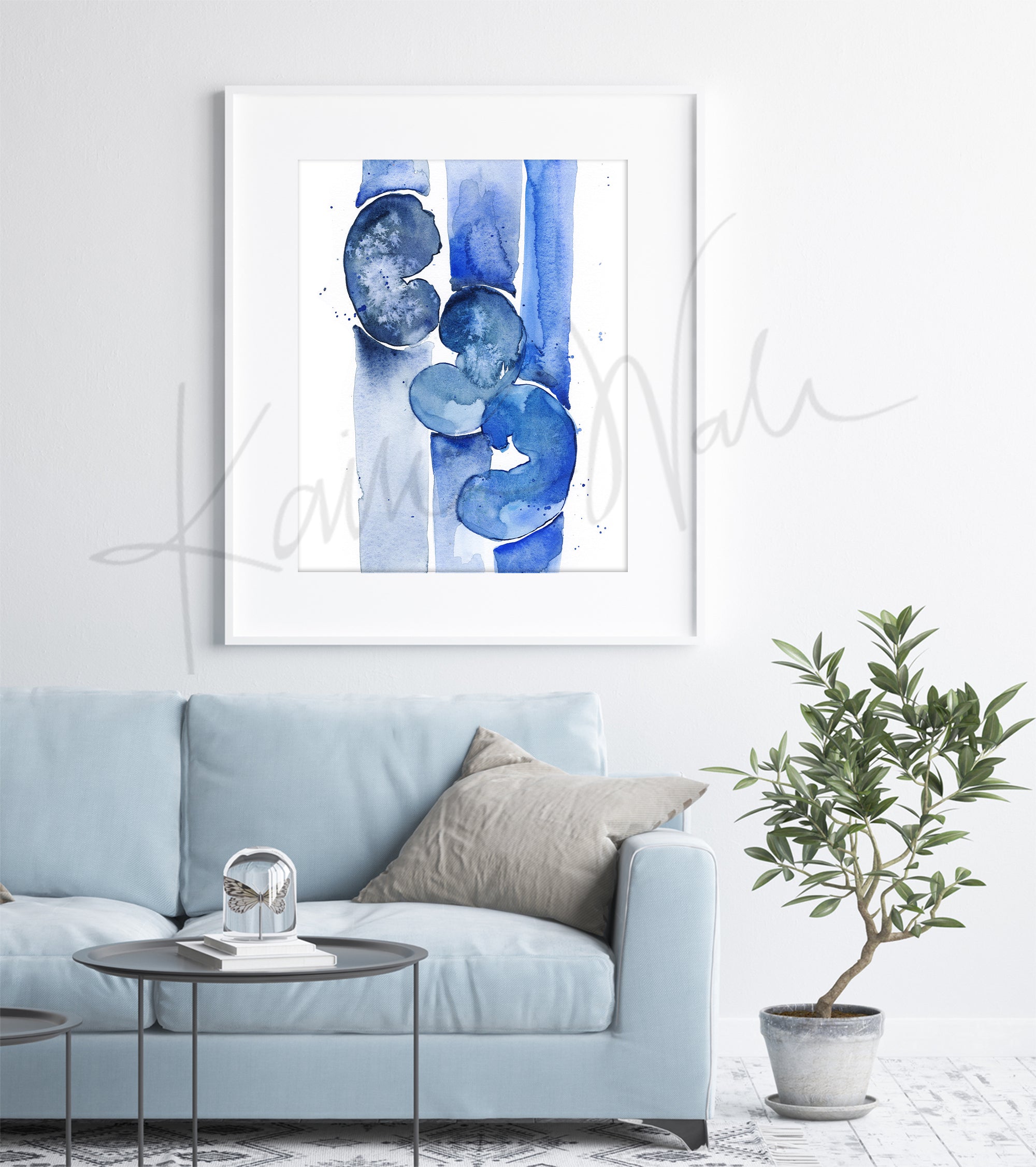 A watercolor painting of three kidneys in blues and navy. It is abstract, but still clearly kidneys to a nephrologist. The anatomy art print is in a black wood frame and hanging above a beige couch with a cushion, like in a living room.