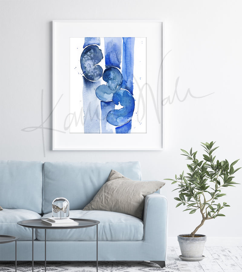 A watercolor painting of three kidneys in blues and navy. It is abstract, but still clearly kidneys to a nephrologist. The anatomy art print is in a black wood frame and hanging above a beige couch with a cushion, like in a living room.